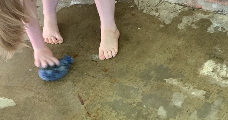 View looking down of the concrete with that's mainly wet. You can see Ada's feet, her hair draping down, and one hand wiping the ground with a crocheted water balloon.