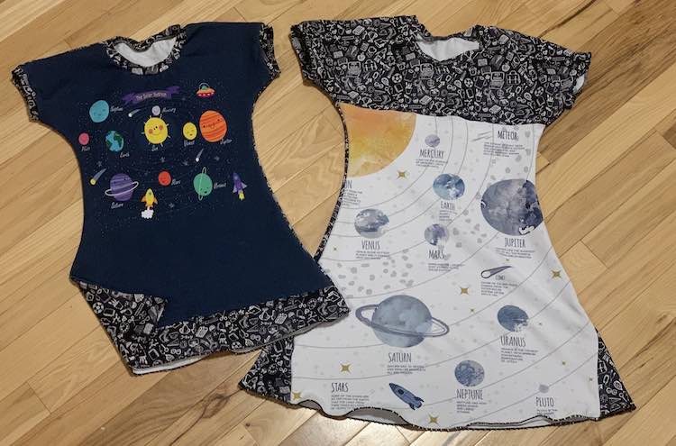 Flat lay of the two dresses side by side. The one on the left are cartoon-ish planets on a navy blue background with the school doodles for the bands along the neckline, cuffs, and along the bottom of the dress. The other dress had a diagram of the planets with text about each one added. The doodle fabric for the bands don't stand out here as the coordinating doodle fabric was added to the bottom sides of the panel along with the top bodice. 