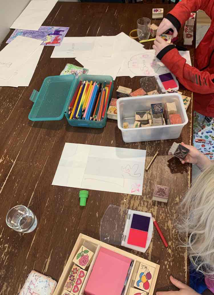 Over view of the girls sitting at the table decorating white paper bags. There's a container of pencil crayons between them and a bunch of stamps scattered around. 