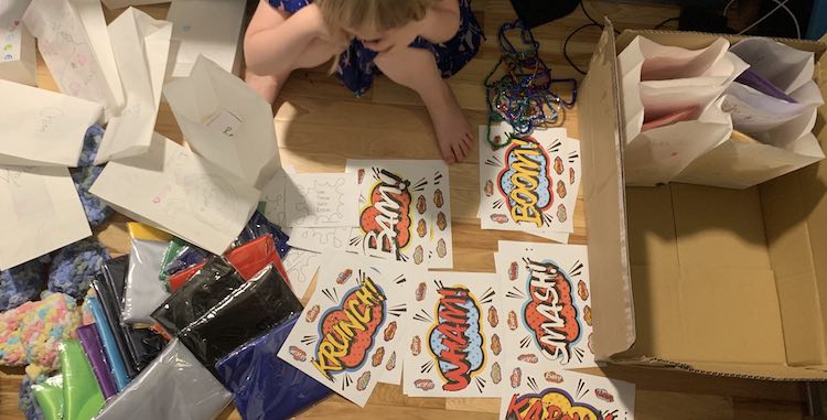 Overview of the treat bags and all it's contents spread out on the floor. Ada is sitting at the top of the photo, the empty bags to the left, a box with four filled bags to the right, and the water balloons, pipe cleaner shapes, capes, and stickers laid out around her.