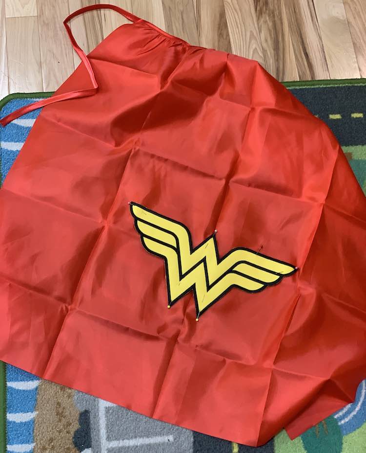 Closeup of the red cape with the black edged yellow Wonder Woman log pinned on. The yellow pins stand out at the top and bottom of the W but there are other smaller ones along the top of the logo too.