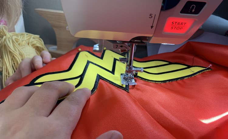 Closeup of the logo on the cape as it's being sewn together in my sewing machine. My left hand holds the fabric while my right hand snapped the photo. To the left you can see Ada's hand, a doll's head, and part of Ada as she watches.