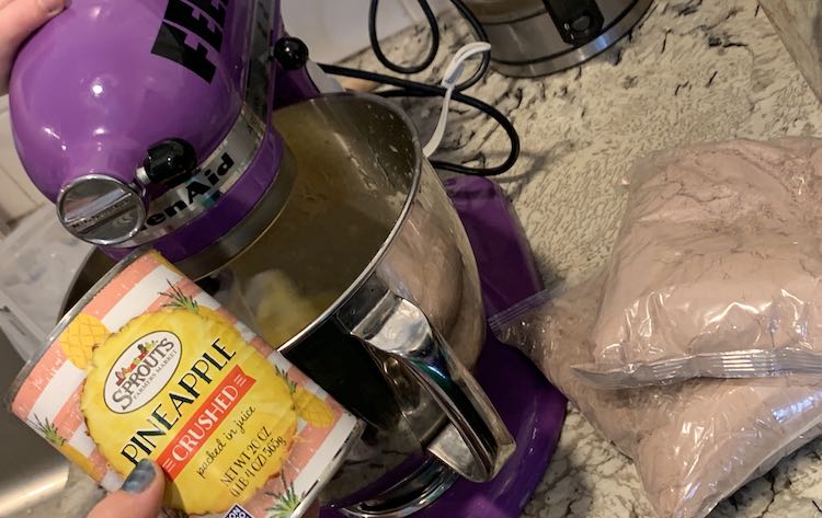 The purple stand mixer is currently beating the contents of the bowl. Ada's hand is resting on top of the mixer while I hold the emptied can of crushed pineapple up for the camera. Off to the right, on the counter, sits the two unopened bags of brownie mix. 