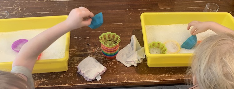 Image shows two yellow rectangular bins with soapy water in it. Between the bins are two white washcloths and a stack of dirty silicone cupcake liners. The back top of the kids' heads are showing and they're currently putting liners in the soapy water. 