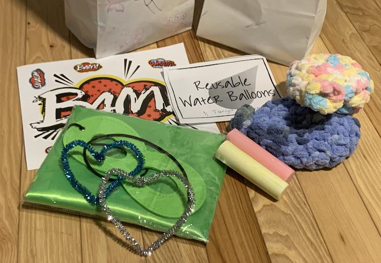 Image showing the contents of the treat bags with two treat bags sitting behind it. One item is a white sheet with comic book Bam, Kaboom, etc stickers. There's also a printed rectangle of paper saying "Reusable