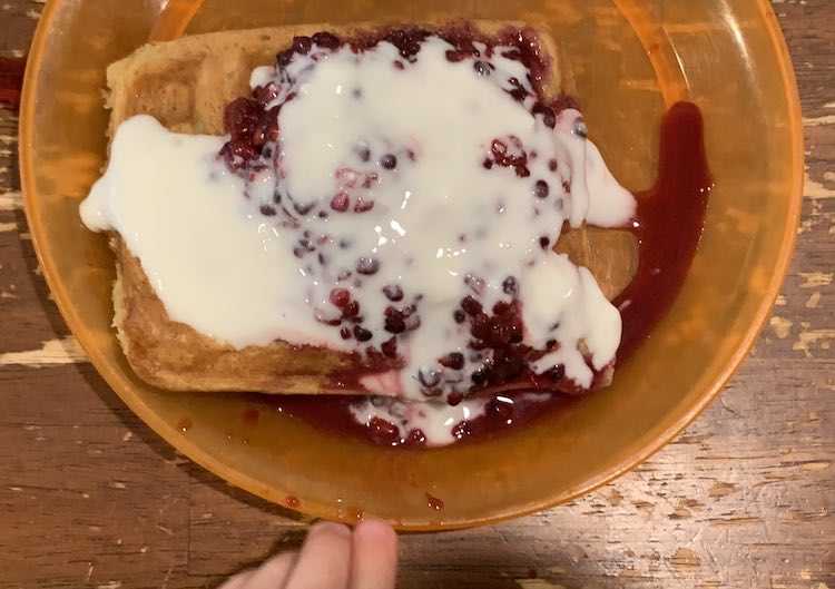 Closeup of an orange plate with little fingers beside it. On the plate is a single waffle with a pile of blackberries and coated in a white sauce. 