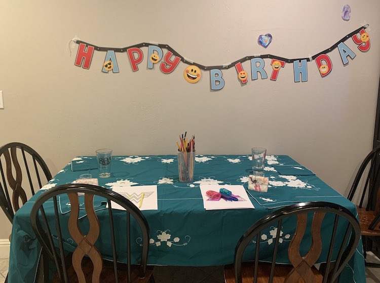 A wooden table covered in a blue/green table cloth with napkins, glasses, papers, and coloring pencils on it. Around the table sits four chairs and above the table is a Happy Birthday banner on the wall.