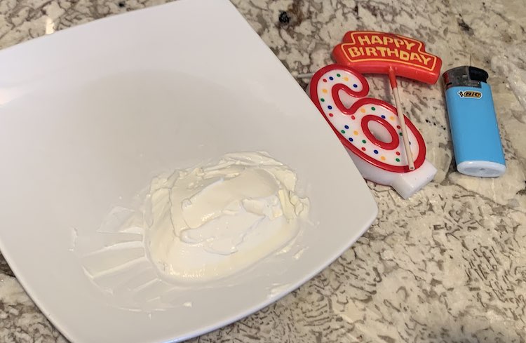 Image shows the kitchen counter with two birthday candles, a lighter, and a square white saucer with a dollop of cream cheese on the side. 