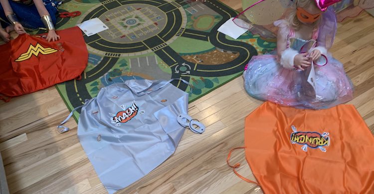 View looking down at the floor of three capes: red, silver, and orange. I quickly finished attaching the stickers to the silver cape while the kids are still crouched in front decorating them.