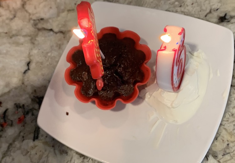 View from above of a brownie cupcake in a red flower silicon liner sitting on a white square plate on the kitchen counter. In front of the brownie sits a number 6 candle, lite, on a pile of cream cheese. 