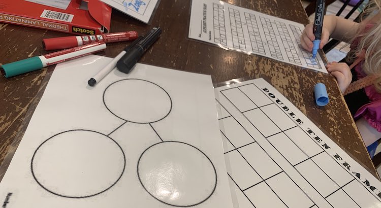 Image shows the two grade one laminated worksheets on the table. In the background Zoey uses a blue chalk marker to trace a letter, on the right side, while the laminating pouches, more markers, and the Zoey page sits on the left side. 