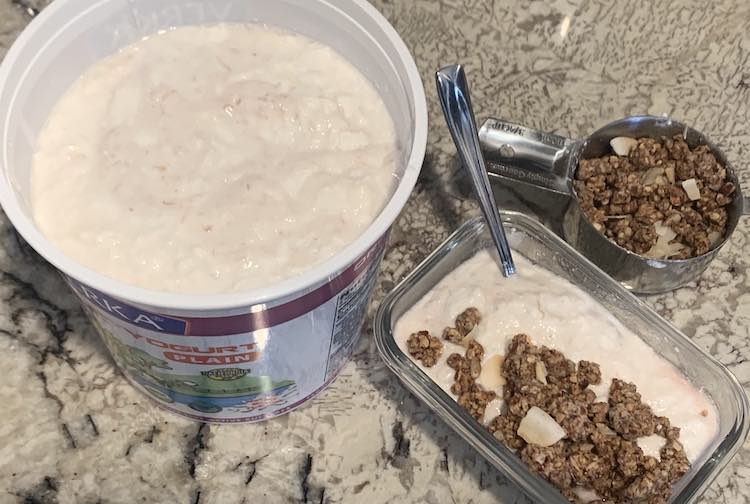 Image shows a 64 oz. container filled with homemade yogurt with dollops of pink-ish yogurt at the top. To the side is a glass rectangular container and a metal spoon sitting in yogurt and granola. Behind it is a measuring cup mostly filled with granola. 