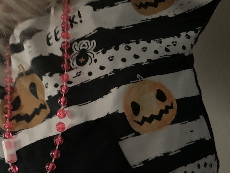 Image shows a closeup of a portion of a shirt mid torso. There's a pink necklace and black kangaroo pocket to the left. Above and beside the pocket sits a little jack-o-lantern that used to be white with black features and now is shaded with an orange fabric marker. 