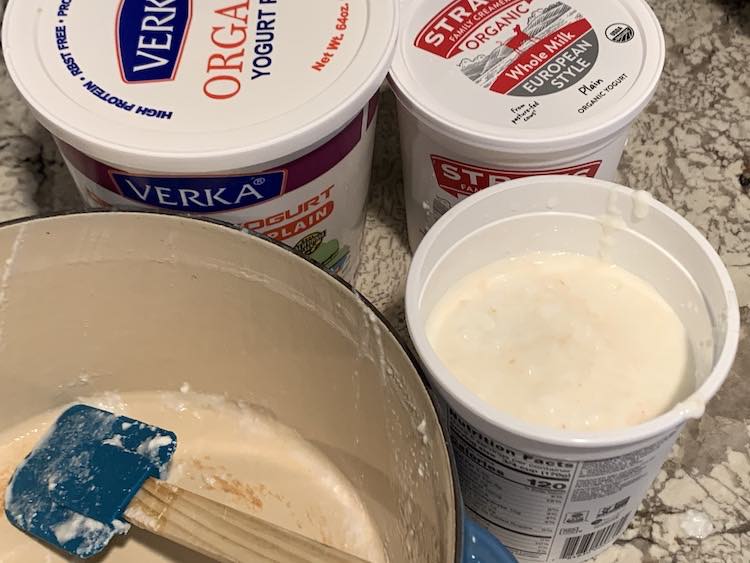 Overview of an emptied Dutch oven showing scald marks in the center and a blue spatula. Behind it sits one large and lidded container of yogurt, one small lidded container of yogurt, and, beside it, one small opened container of yogurt with brown flecks inside.