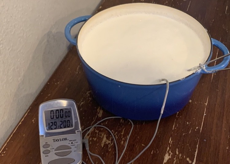 Image shows the Dutch oven sitting on the table (potholder hidden underneath) with the whisk and thermometer tip still in the milk. The temperature gauge shows it's at 129°F.