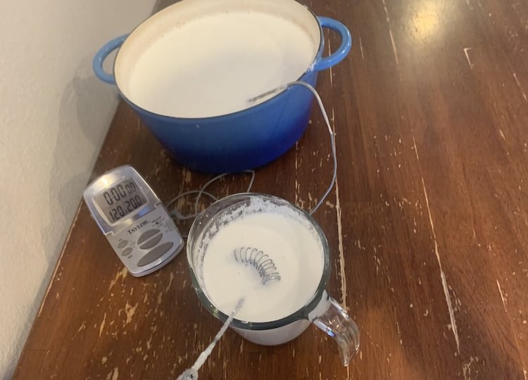 Image shows the Dutch oven on the table with the thermometer tip still in it. The temperature is shown to be 120°F. In front of both items sits a one cup jug filled with milk and a metal whisk.