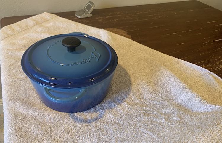 Image shows a white towel laying on the table with the lidded Dutch oven sitting in the center of it. Off in the back is the thermometer without the tip attached and, part, of the potholder.