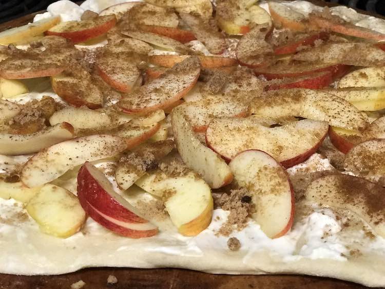Image shows a closeup of a beige pizza crust with white cream cheese, sliced apples, topped with brown sugar and cinnamon.