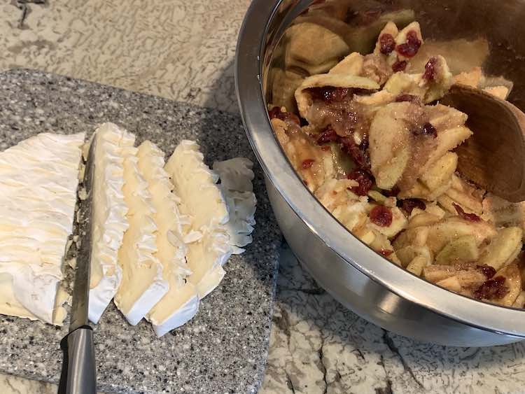 Image shows a metal bowl filled with sliced and peeled apples, craisins, and seasonings. Beside it sits a small marbled cutting board with sliced brie cheese with the rind on. 