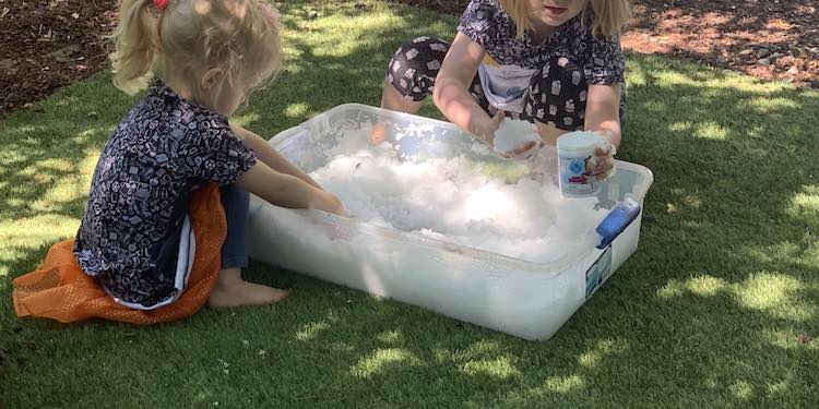 Closeup of a bin of fake snow with Ada crouched behind it and Zoey sitting beside it. Both are wearing their matching dresses. 