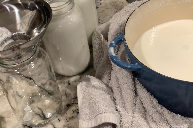 Image shows my blue Dutch Oven with yogurt inside sitting on a white towel. Off to the side sits two filled glass canning jars and an empty one, in front, with a metal wide mouth funnel sitting in the top. 