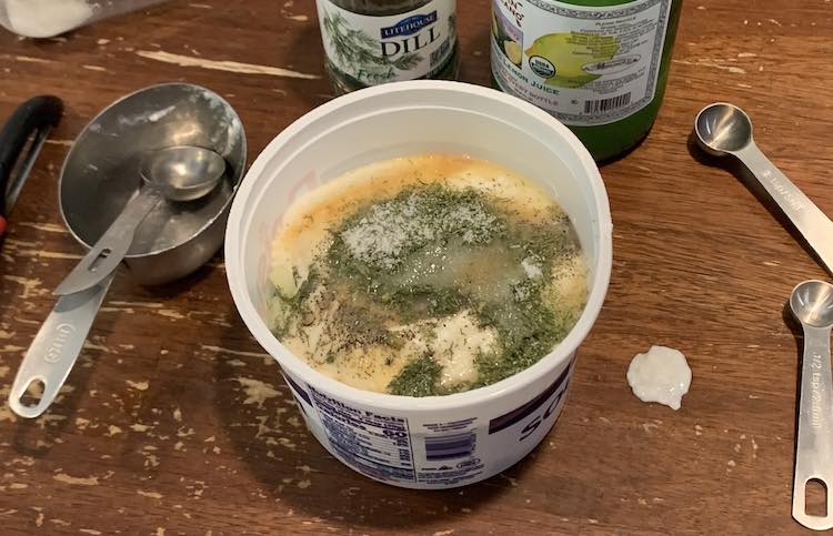 Image shows a plastic container filled with green (dill), white (sugar and yogurt), and beige (garlic). Off to the sides are a vegetable peeler, measuring spoons, and a measuring cup. Behind it sits a lemon juice bottle and a dried dill container.