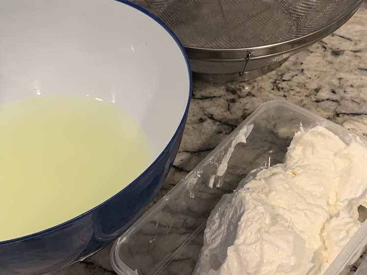 Image shows a small glass rectangular container, lower left, holding a mound of white cream cheese looking stuff. Beside it and back a bit sits a blue plastic bowl with yellowy liquid inside the white interior. Behind both sits a metal bowl-like strainer. 