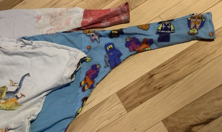 Three knit shirts laying on a wooden floor. You can only see a single sleeve of all three. The top is a white short sleeved dinosaur top with a small triangle right before the band at the sleeve. The next two shirts have a long sleeve. The middle blue one has a seam around the armpit while the bottom hearted one has three fabrics showing with the first seam halfway down the arm and another closer to the end where a wide band was added.