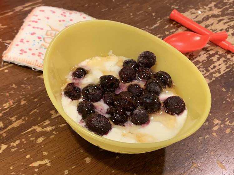 Image shows a plastic yellow bowl half filled with white yogurt topped with frozen blue-black blueberries and drizzled with yellow honey. Behind the bowl to the left sits Zoey's fabric name-tag and to the right lays a separated plastic cereal spoon.