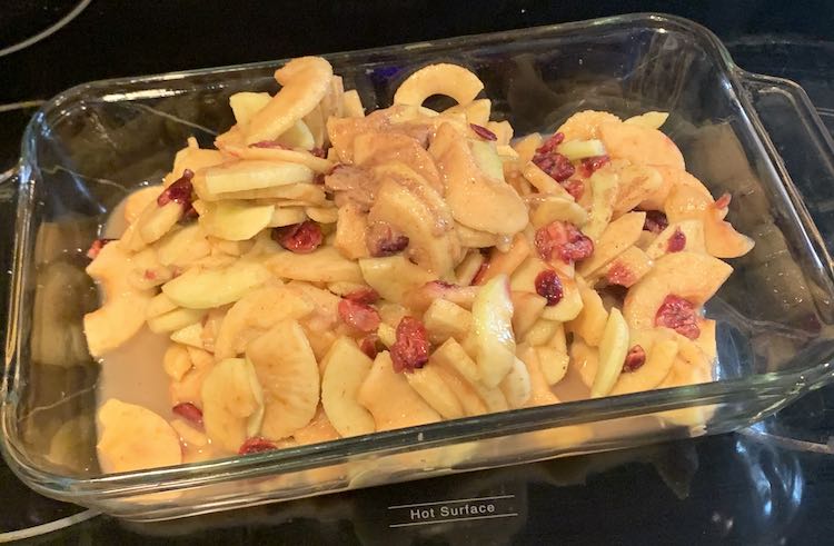 Image shows a closeup of a clear casserole dish sitting on a stovetop. In the dish is a pile of apple slices, craisins, and a brown sugar syrup clumped in places and a pool below. 