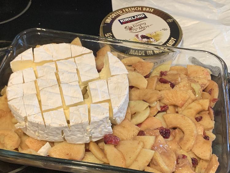 Image shows the glass casserole dish filled with a flat layer of apples, craisins, and sugar syrup. On the left sits a partially coming apart wheel of brie. Behind the casserole dish sits the flattened brie wrapper and the brie container from Costco.