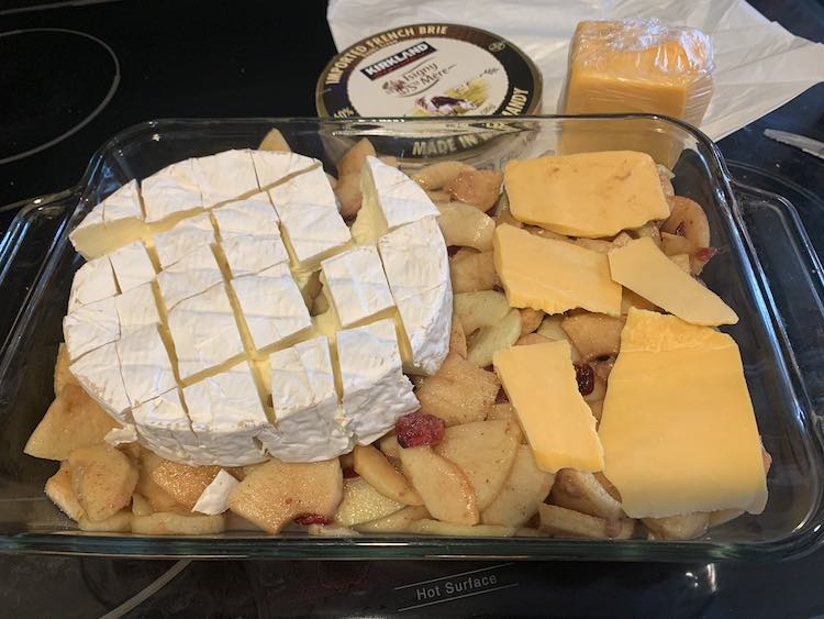 Image shows the casserole dish half filled with the apple pie filling. On the left sits the brie and on the right are five mismatched slices of orange cheddar cheese. Behind the casserole dish sits the brie wrapper and the wrapped leftover cheddar cheese. 