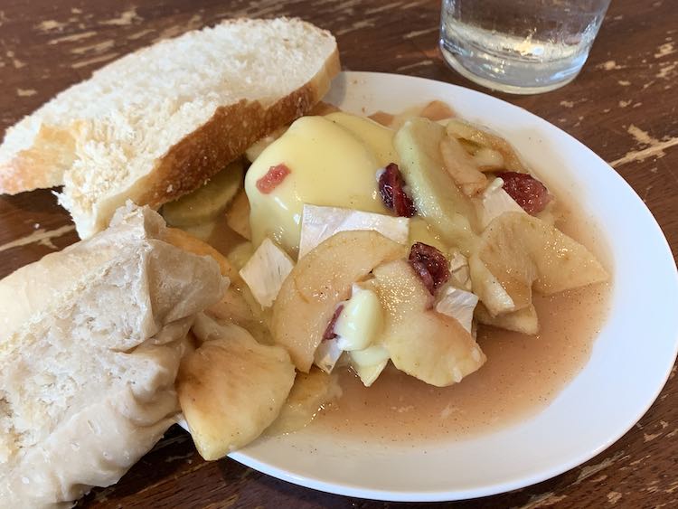 Image shows a plate with a pile of brie coated apples and crasins on it sitting in syrupy sauce. Leaned against the side of the plate are two slices of bread; in the back sits a dried slice of store bought bread while in the front sits a soft dumpling-like slice. 