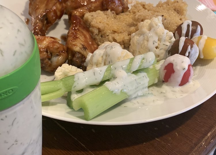 Image shows part of the OXO salad dressing container in the foreground. Behind it sits a plate with chicken wings, quinoa, and veggies drizzled with the ranch dressing.