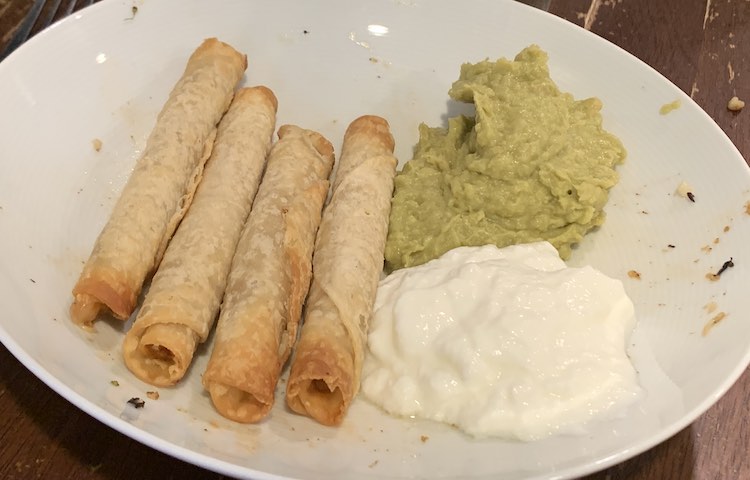 Image shows a white plate on a table with four browned taquitos laying in a row next to a pile of white yogurt and green avocado hummus.