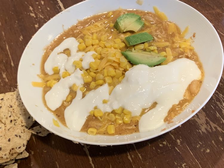 Image shows a white bowl on a wood table with corn tortillas sitting next to the bowl. In the bowl sits a brown-ish soup hidden under corn, shredded cheese, avocados, and white yogurt.  