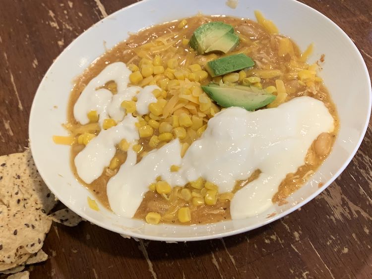 Image shows a white bowl on a wood table with corn tortillas sitting next to the bowl. In the bowl sits a brown-ish soup hidden under corn, shredded cheese, avocados, and white yogurt.