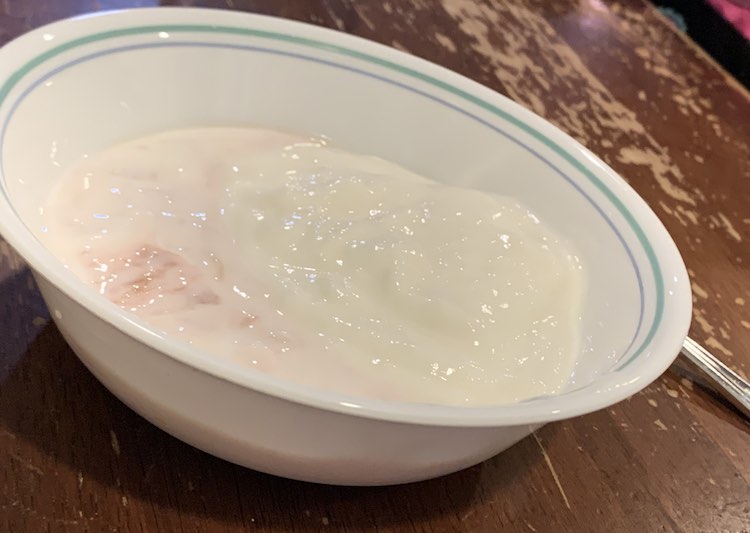 Image shows a white bowl with a portion of a metal spoon showing in the back. There's a moon shaped pink-ish yogurt on the left with a couple chunks of rhubarb. The rest of the bowl is filled with a mound of white yogurt. 