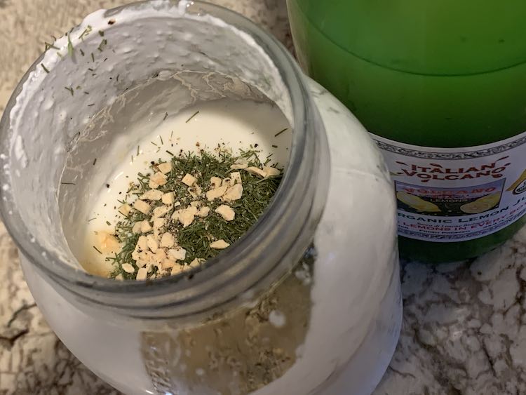 Image is taken slightly from above of an opened canning jar showing a bit of yellow liquid, green flakes (dill), and chunks of brown-ish white (dehydrated garlic). In the background is a bottle of lemon juice from Costco. 