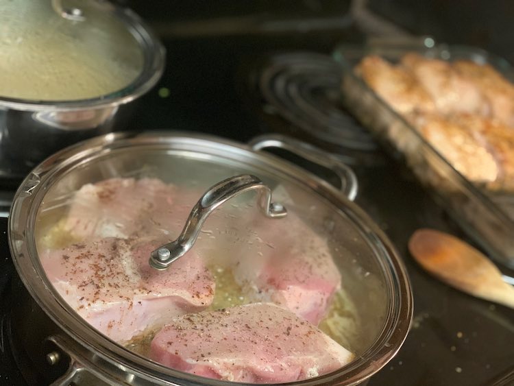 Image shows a clear lidded metal frying pan with some raw looking seasoned, black speckled, pork chops frying in butter. In the background is a lidded pot and a casserole dish already half filled with seared pork chops.