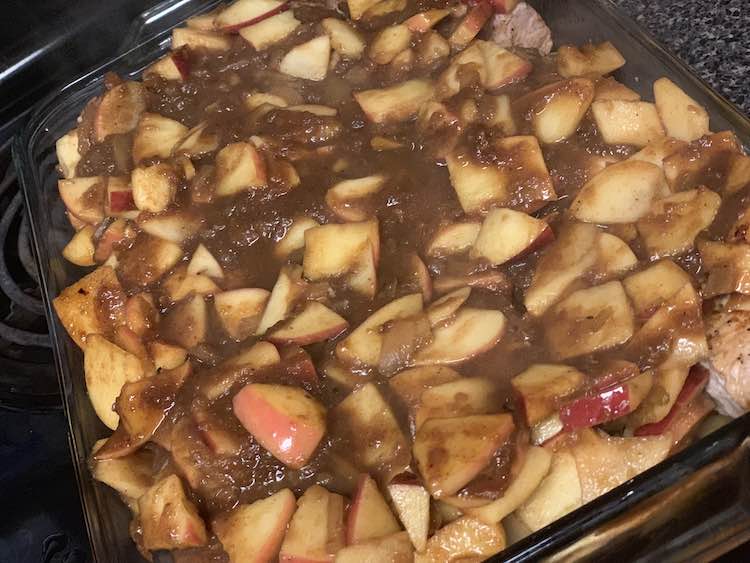 Image shows a closeup of the glass casserole dish filled withe chunks of apples and brown sauce. The pork chops are mostly hidden underneath.