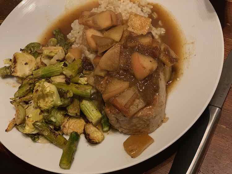 Image shows a white plate with a steak knife off to the side. On the plate is a pile of roasted diced Brussel sprouts and asparagus. Beside it is a pile of rice topped with a pork chop and apple topping. Around the food you can see the apple butter sauce. 