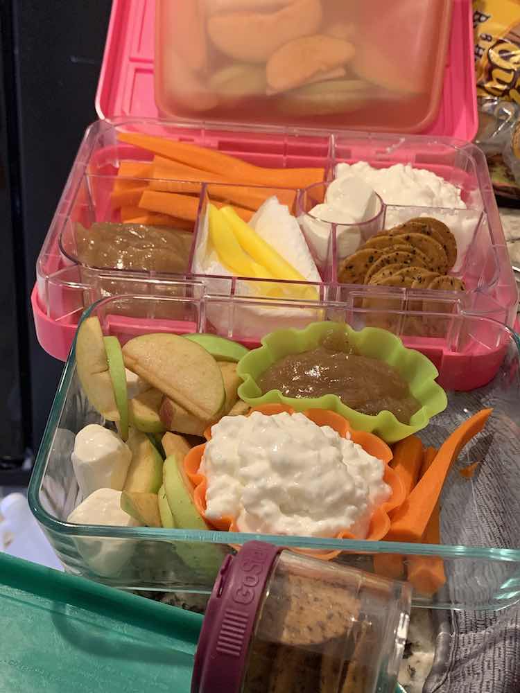 Image shows an opened lunchbox with an apple filled stasher bag leaned against the lid and a glass container, with similar stuff, in front of it. There's a small plastic container of crackers in the front. The dried, wet, and sauces are separated by the lunchbox sections of silicon cupcake liners. 