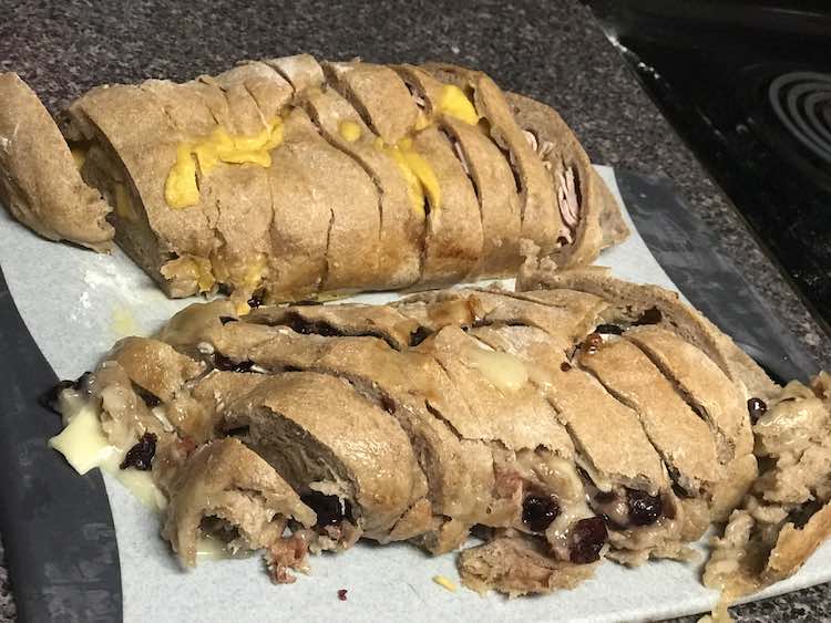 The image shows parchment paper with two loaves sitting on it. Both loaves are sliced. The back loaf has cheddar cheese on it and some deli meat showing at either end. The loaf in the front shows craisins and brie oozing out from the sliced sections.