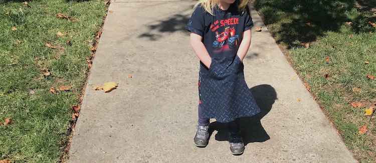 View of Ada's monster truck dress with matching tire tread fabric. Ada's standing straight with her hands in her pocket enjoying the kangaroo pocket in the skirt. 