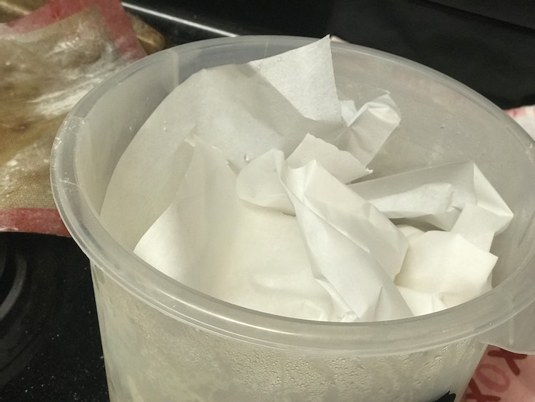 Image shows a closeup of the top side of the plastic breadin5 bin with parchment crinkled up as if it was a gift basket. In the background sits a flour covered silpat sheet and a kitchen towel. 
