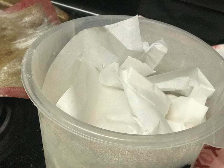 Image shows a closeup of the top side of the plastic breadin5 bin with parchment crinkled up as if it was a gift basket. In the background sits a flour covered silpat sheet and a kitchen towel.