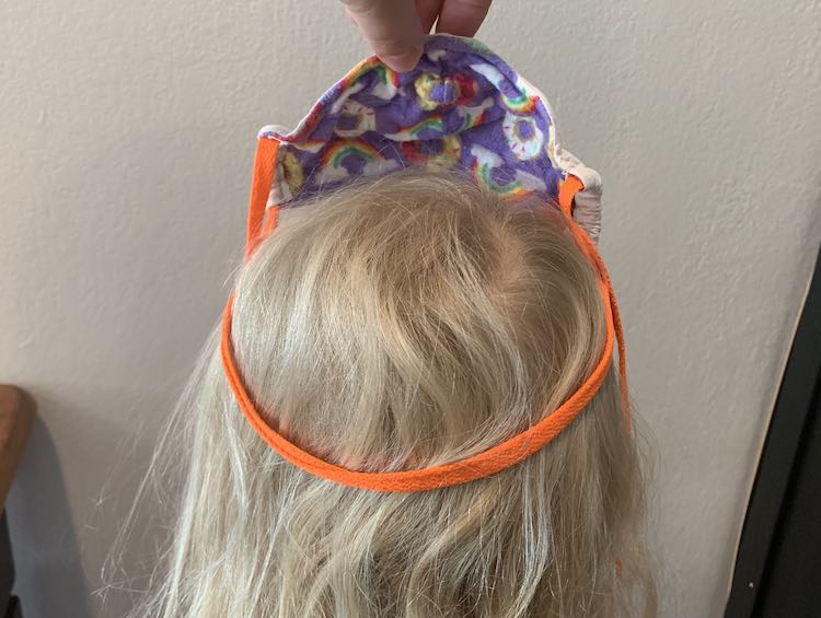 Image shows me holding the mask on Zoey's head like a crown. The lower loop goes around her head while the two ends of the shoelaces droop down from the two upper corners of the mask. 