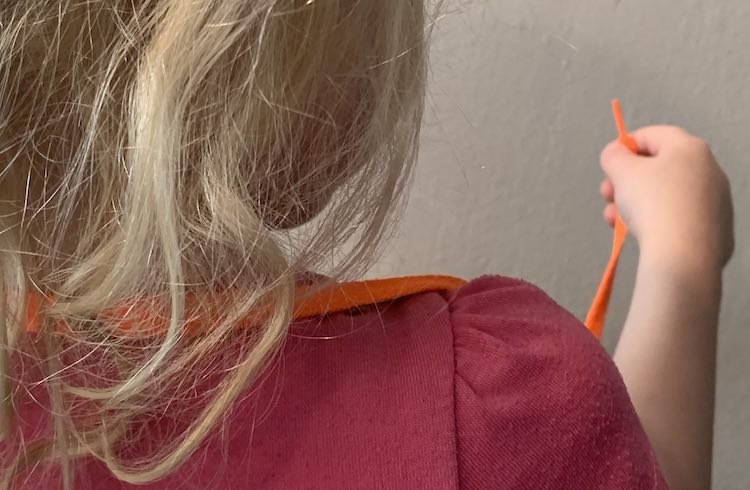 View of the back of Zoey's head and a shoulder. The orange shoelace is looped around her neck with the mask hidden in front of her. Her right hand is holding an end of the shoelace. 