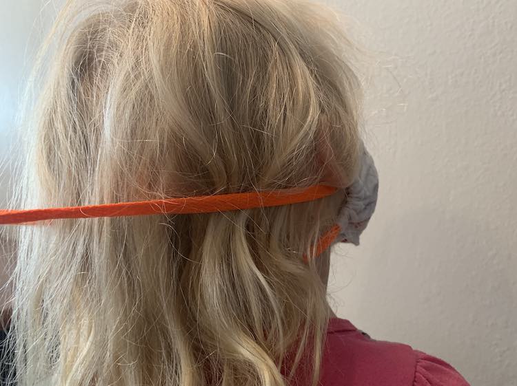Image is taken from the back. You can see a part of the lower shoelace as it extends from the lower channel and disappearing under Zoey's hair. The upper shoelace is extending from the top of the mask and goes under Zoey's ear before stretching out. 